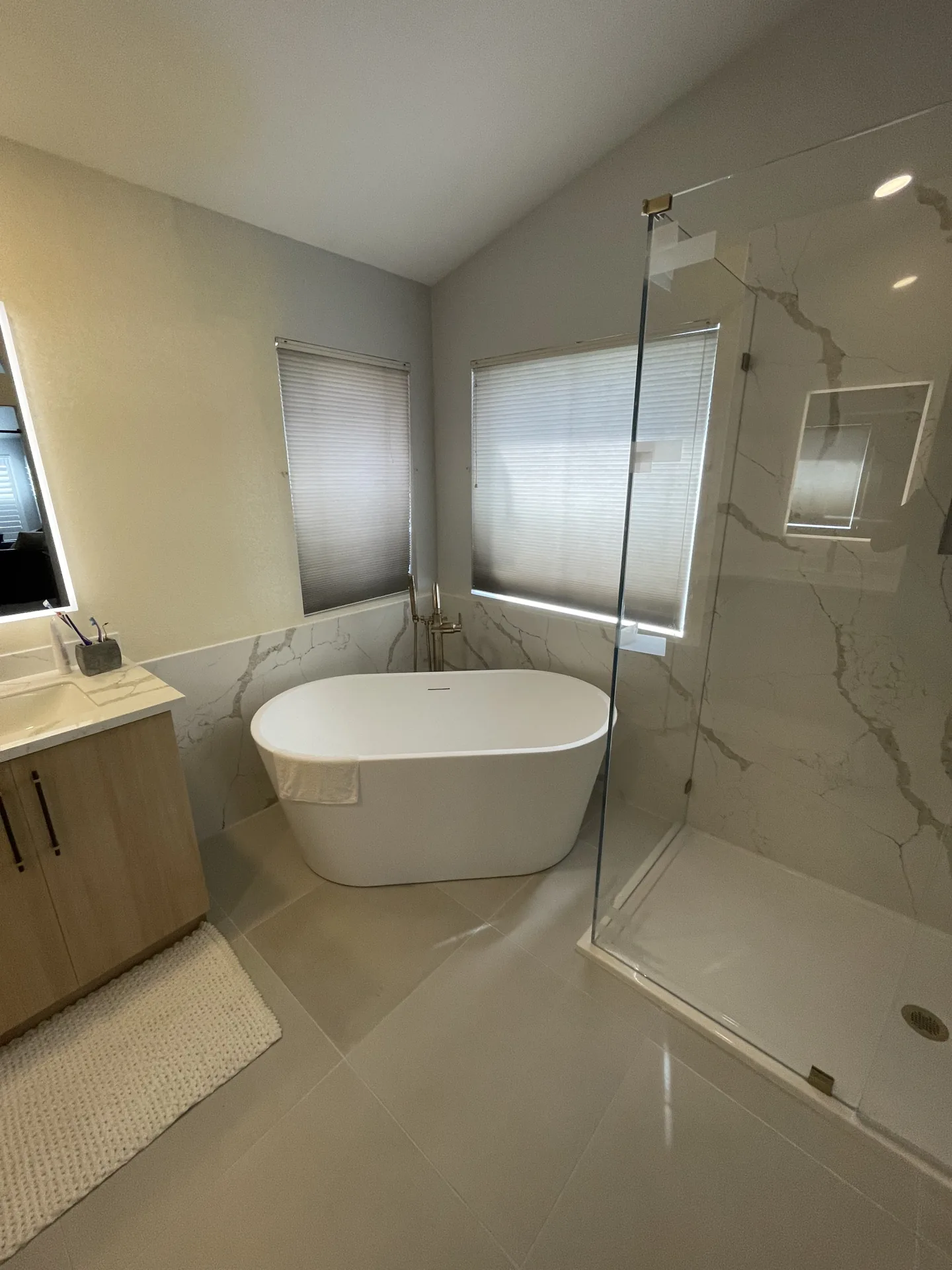 Bathroom remodel showcasing material selections — marble-look quartz walls, a freestanding tub, light wood vanity, and brass fixtures
